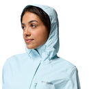 A woman wears a light blue Columbia Womens Pouring Adventure III Waterproof Hiking Jacket with the hood up, looking to the side against a plain white background.