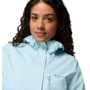 A woman with curly dark hair, facing forward and smiling slightly, wears a light blue Columbia Womens Pouring Adventure III Waterproof Hiking Jacket with a hood. The Columbia logo is visible on the chest against a white background.