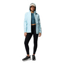 A woman faces forward against a white background, wearing a Columbia Womens Pouring Adventure III Waterproof Hiking Jacket in light blue, a dark blue crop top, black leggings, black sneakers, and a white cap.