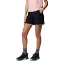 A person wears a light pink sleeveless button-up shirt, Columbia Womens Silver Ridge Elite Hiking Shorts in black, gray socks, and black athletic shoes with light green soles, posing against a white background.