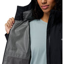 A person wearing a light blue shirt holds open a black Columbia Womens Trailborne 2.5L Waterproof Shell Jacket, showing its gray patterned lining and small mesh pocket. Only the lower half of their face and torso are visible.