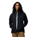 A woman with long curly hair stands with hands in pockets, wearing the Columbia Womens Trailborne 2.5L Waterproof Shell Jacket in black over a light blue shirt and tan pants, against a plain white background.