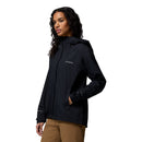A woman with long curly hair wears a black Columbia Womens Trailborne 2.5L Waterproof Shell Jacket and tan pants, standing against a plain white background.