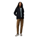 A woman with long, curly hair stands facing forward against a plain white background, wearing the Columbia Womens Trailborne 2.5L Waterproof Shell Jacket in black, paired with a light blue top, brown pants, and beige sneakers. Her hands are in her jacket pockets.
