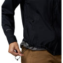 A person tightens the drawstring at the hem of a black Columbia Womens Trailborne 2.5L Waterproof Shell Jacket over tan pants. The jacket features a zipper and a hook-and-loop strap on the sleeve.