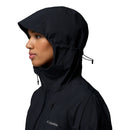 A woman is shown in profile against a white background wearing the Columbia Womens Trailborne 2.5L Waterproof Shell Jacket in black, with the hood up and cinched tightly with the drawstring.
