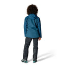 A person with curly hair, wearing the Rab Womens Latok Mountain GORE-TEX Pro Waterproof Jacket in blue, black pants, and teal-accented shoes, stands facing away against a plain white background.
