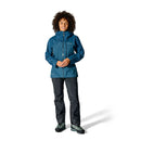 A woman stands facing forward and smiling against a plain white background, wearing the Rab Womens Latok Mountain GORE-TEX Pro Waterproof Jacket in blue, paired with black pants and green hiking boots.