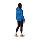 A woman stands facing away, wearing a Rab Womens Microlight Alpine Hoody in blue styled as a puffer, paired with black leggings, beige sneakers, and striped socks. The background is white.