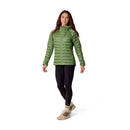 A woman wearing the Rab Womens Microlight Alpine Hoody in green, paired with black leggings, beige hiking boots, and striped socks, stands and smiles against a plain white background.