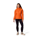 A person stands facing forward, wearing a Rab Womens Microlight Alpine Hoody in orange, styled as a puffer jacket, paired with black leggings and beige hiking shoes, against a plain white background.