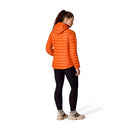 A person stands facing away, wearing the Rab Womens Microlight Alpine Hoody in orange, paired with black leggings and beige sneakers, against a plain white background.