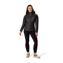 A woman stands smiling against a white background, wearing the Rab Womens Microlight Alpine Hoody, black leggings, and beige hiking boots.