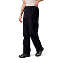 A person stands against a white background wearing Rab Womens Namche Hike GORE-TEX Pants in black, paired with a light short-sleeve shirt and hiking shoes. Their left arm, which is tattooed, rests relaxed at their side.