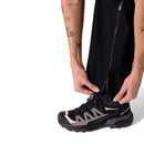 A person with tattoos on their arm is zipping the side of their black Rab Womens Namche Hike GORE-TEX Pants over a black and beige Salomon sneaker.