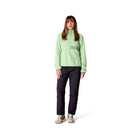 A woman stands facing forward, wearing a light green Rab Womens Ramshaw Pull-On Fleece, dark pants, and beige sneakers, against a plain white background.