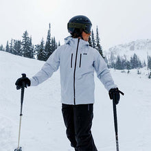 Man standing while skiing in a light grey Helly Hansen ski jacket with black pants on a white snowy mountainside.