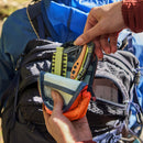 Outdoors, a person opens a Leatherman® Tool Pouch attached to their backpack, showing a Leatherman multi-tool and an orange folding knife inside. Another backpack with weather-resistant zippers is visible in the background.