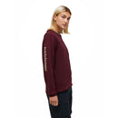 A woman with shoulder-length blond hair wears the Peak Performance Womens Trail Polartec Power Grid Crew Fleece, a maroon long-sleeve pullover featuring branding on the left sleeve, paired with dark pants and standing against a plain white background.
