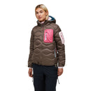 A person is wearing the Peak Performance Womens Helium Utility Down Hood Jacket in brown with pink pockets and dark pants, standing facing forward against a white background.
