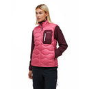 A person with blonde hair wears the Peak Performance Womens Helium Utility Down Vest in pink, featuring a maroon chest pocket, layered over a maroon long-sleeve shirt and paired with black pants, standing against a white background.