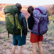 Two people with large hiking backpacks standing in a desert landscape at sunset.