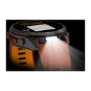 Garmin fēnix 9 Pro inReach with an orange strap shown displaying a fitness screen. A bright LED light on the side of the watch is switched on, casting a forward beam. The display appears black against a dark background. This multisport GPS smartwatch offers advanced features ideal for night workouts and outdoor adventures.