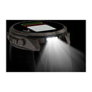 A close-up of the Garmin fēnix 9 Pro inReach multisport GPS smartwatch with its built-in flashlight illuminated, casting a bright white beam forward in a dark environment. The watch display shows battery and activity icons, showcasing the advanced features of the fēnix 9 Pro inReach, including satellite connectivity for precise tracking on any adventure.