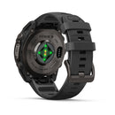 A dark gray Garmin fēnix 9 multisport GPS smartwatch is shown from the back, displaying its glowing green and orange sensor lights. It features a silicone wristband and a metal buckle.