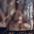 A Leatherman Rev Multitool with an open locking blade stands upright in a dark wooden surface. The blurred background shows earth tones and abstract shapes.