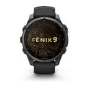 A black Garmin fēnix 9 multisport GPS smartwatch featuring a round AMOLED display with “FENIX 9” displayed over a gold topographic map design. The watch includes side buttons and a sleek black wristband.