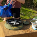 A person pours water from a blue container into a Sea to Summit Camp Kitchen Clean Up Kit (6-Piece) portable sink on a table outdoors, preparing their camp dishwashing setup. Soap and a cooking pot are nearby, making campsite kitchen clean-up quick and convenient.