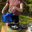 A person pours water from a blue lightweight container into a Sea to Summit Folding Bucket (10L) outdoors, with camping gear and greenery visible in the background.
